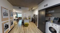 Located at 1900 Rio Grande Street in Austin, Texas, this single-floor unit presents a contemporary industrial aesthetic defined by exposed concrete ceilings, light wood-style flooring, and crisp white walls throughout. The open-concept kitchen serves as a central hub of the living space, featuring stainless steel appliances—including a full-size refrigerator, electric range, and microwave—paired with dark lower cabinetry, white upper cabinets, and a kitchen island with additional prep space. Track lighting overhead provides practical illumination across the cooking and dining areas.

The living area benefits from large windows that bring in generous natural light and offer elevated views of the surrounding Austin cityscape, complemented by exposed concrete columns that reinforce the unit's industrial character. Multiple bedrooms are present, each with individual windows providing outdoor views, wood-style flooring, and space for a bed and desk setup. The bathroom includes a full tub-shower combination and a dark-finished vanity with tile flooring. A dedicated in-unit laundry area with a washer and dryer adds everyday convenience. The layout, as seen in the floor plan, accommodates multiple occupants comfortably across well-defined living zones.
