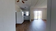 This single-story space features an open-concept layout that seamlessly connects the kitchen and living area, creating a cohesive and functional environment. Dark hardwood floors run throughout, providing a warm contrast to the predominantly white interior. The kitchen is equipped with white cabinetry, a refrigerator, a range, and a sink positioned beneath a window that allows natural light to filter in. Dark countertops add depth to the kitchen's clean aesthetic. A ceiling fan is mounted overhead, enhancing airflow across the open layout. The living area extends naturally from the kitchen, offering ample room for furnishings. A sliding glass door at the far end of the living space serves as a prominent architectural feature, flooding the interior with abundant natural light and providing direct access to an outdoor area. The textured ceiling adds subtle visual interest throughout. Overall, the space presents a straightforward, functional design with an open feel, benefiting greatly from the natural light provided by both the kitchen window and the expansive sliding glass door.