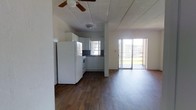 This single-story space features an open-concept layout that seamlessly connects the kitchen and living area, creating a cohesive and functional environment. Dark hardwood floors run throughout, providing a warm contrast to the predominantly white interior. The kitchen is equipped with white cabinetry, a refrigerator, a range, and a sink positioned beneath a window that allows natural light to filter in. Dark countertops add depth to the kitchen's clean aesthetic. A ceiling fan is mounted overhead, enhancing airflow across the open layout. The living area extends naturally from the kitchen, offering ample room for furnishings. A sliding glass door at the far end of the living space serves as a prominent architectural feature, flooding the interior with abundant natural light and providing direct access to an outdoor area. The textured ceiling adds subtle visual interest throughout. Overall, the space presents a straightforward, functional design with an open feel, benefiting greatly from the natural light provided by both the kitchen window and the expansive sliding glass door.