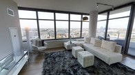 This elegant high-rise residence offers a sleek, modern living experience defined by its most striking feature: expansive floor-to-ceiling windows that wrap around the living area, flooding the space with natural light and framing breathtaking panoramic views of the city skyline below. The open-plan layout is thoughtfully designed to maximize the connection between the interior and the urban landscape beyond.

The living area is appointed with a refined, all-white furniture ensemble, including a generous sectional sofa, tufted ottoman cubes, and a sculptural accent chair, all anchored by a plush dark shag area rug that adds warmth and contrast to the bright palette. A statement arc floor lamp with a metallic shade adds a bold, contemporary touch, while hardwood flooring runs throughout, lending a clean and cohesive aesthetic.

The architecture emphasizes clean lines and a minimalist sensibility, with a neutral color scheme that keeps the focus on the stunning cityscape visible from nearly every angle. Track lighting overhead provides versatile illumination to complement the abundant natural light. The overall ambiance is one of urban sophistication — bright, airy, and meticulously curated for modern living.