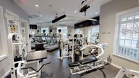 This fitness center is a comprehensively outfitted facility designed to support a full range of workout routines. The space is finished with durable speckled rubber flooring throughout, providing both safety and functionality for all types of exercise. Neutral gray walls and recessed lighting create a clean, focused environment conducive to training.

The cardio section features multiple treadmills, elliptical machines, and stationary bikes, all arranged along a wall of floor-to-ceiling mirrors that visually expand the space and allow users to monitor their form. Multiple flat-screen televisions are mounted at eye level throughout the room, offering entertainment during workouts.

The strength-training area is well-stocked with a variety of Paramount and other branded cable machines, weight benches, and multi-functional gym stations, catering to both upper and lower body training. Natural light streams in through several white-framed windows fitted with horizontal blinds, keeping the space bright and inviting.

Framed motivational artwork decorates the walls, adding a polished, purposeful aesthetic. A glass-paneled door provides access to an adjoining area, enhancing the sense of connectivity within the facility. The ceiling fan and ventilation system contribute to a comfortable workout environment. Overall, this fitness center offers a professional-grade experience in a well-maintained, thoughtfully designed setting.