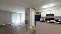 This single-floor residence offers a cohesive and functional layout finished with warm wood-look flooring throughout. The open kitchen is a standout feature, equipped with crisp white cabinetry, dark granite-style countertops, black appliances including a refrigerator and range, and a peninsula that provides both additional prep space and a natural divide between the kitchen and living areas. A flush-mount ceiling light brightens the kitchen, which also features a textured ceiling consistent throughout the unit. The living area is generously sized, with neutral warm-gray walls, white baseboards, and access to a sliding glass door that lets in natural light. A separate entry hallway with wood-look tile flooring provides a smooth transition into the main living spaces. The bathroom is thoughtfully designed with a white vanity cabinet, dark countertop, a tiled tub-shower combination, and wood-look flooring. The bedrooms are well-proportioned with warm neutral walls, wood-look floors, and natural light from blinded windows. At least one bedroom features a spacious double-door closet with a full-width hanging rod, offering excellent storage. The textured ceilings and consistent neutral palette lend the entire unit a clean, cohesive aesthetic.