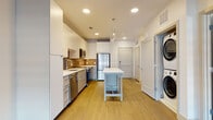 This thoughtfully designed single-floor residence combines modern finishes with functional living spaces throughout. The kitchen serves as the heart of the home, showcasing sleek white cabinetry extending to the ceiling, a warm tile backsplash, and a full suite of stainless steel appliances including a French door refrigerator, range with over-the-range microwave, and dishwasher. White quartz countertops provide ample prep space, complemented by a central island with a clean white top, ideal for casual dining or additional workspace. Warm-toned luxury vinyl plank flooring runs throughout the kitchen and common areas, adding a welcoming feel. Recessed lighting and a pendant fixture illuminate the space with a bright, contemporary ambiance. A stacked washer and dryer unit is conveniently integrated near the entryway, adding to the home's practicality. The bedroom offers plush carpeting, neutral toned walls, and a wide window that invites natural light while framing urban city views. The bathroom is finished with a single-sink vanity featuring a white quartz countertop, taupe cabinetry, a large mirror, and a glass-enclosed shower, all carried out in a clean, neutral palette. Together, these spaces create a cohesive, modern living environment.