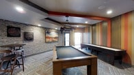 This single-floor recreational space is thoughtfully designed to offer an engaging and social atmosphere for guests and residents alike. The room immediately draws attention with its eclectic mix of design elements — exposed brick walls on one side contrast beautifully with bold, vertically striped accent walls in warm tones of orange, red, and gold on the other. A coffered ceiling with dark accent beams and recessed lighting adds architectural depth, while a striking geometric pendant light fixture serves as a focal point above the central pool table. The full-size pool table with a slate-grey felt surface anchors the space and invites friendly competition. Adjacent to it, a regulation shuffleboard table offers additional entertainment options for guests. Patterned carpet tiles in muted, earthy tones ground the room and complement the warm, contemporary color palette. Natural light filters in through large windows near the entrance, brightening the space and creating an inviting ambiance. Curated wall art, including a vivid landscape painting on the brick wall, adds a finishing touch of personality. Overall, this game room strikes a balance between energetic and comfortable, making it an ideal gathering space for leisure and entertainment.