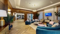 Located at 6290 SW Eighth St in West Miami, Florida, this single-floor leasing office presents a refined and welcoming environment designed to impress. The space is anchored by striking herringbone-patterned wood-look tile flooring that flows seamlessly throughout, complemented by rich, high-gloss wood wall paneling that lends warmth and sophistication to every area.

The main lounge area serves as the centerpiece of the space, featuring curved teal velvet sofas arranged around a round coffee table atop a plush area rug. A statement cluster chandelier with globe pendants illuminates the seating area, while a wall-mounted flat-screen TV and textured accent wall add visual interest. Lush indoor plants bring a touch of nature indoors, enhancing the inviting atmosphere.

The entry foyer greets visitors with a glass-top pedestal table beneath the same elegant chandelier, with floor-to-ceiling sheer drapery framing views to the exterior. Recessed tray ceilings throughout add architectural depth and a sense of volume to the interior.

The corridor connecting the lobby to the elevator bank is lined with glossy wood-clad columns and warm sconce lighting, creating a polished transitional space. Stainless steel elevator doors reflect the refined finishes found throughout. A dedicated mailroom with an expansive bank of individual mail compartments and wood-look flooring rounds out this well-appointed leasing facility.