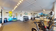 This fitness center offers a comprehensive workout environment designed to support a wide range of exercise routines. The space is laid out across a single floor and is illuminated by track lighting mounted along exposed ceiling beams, creating a bright and energizing atmosphere. Large windows along one wall invite natural light and provide views of the surrounding exterior, enhancing the open feel of the room.

The cardio zone is well-appointed with treadmills, an elliptical, and Echelon spin bikes, each equipped with individual display screens for interactive workout sessions. A large flat-screen TV is mounted on the wall alongside a full-length mirror that visually expands the space and allows users to monitor their form during exercise.

The strength training area features a multi-station cable machine, a tiered dumbbell rack stocked with a wide range of free weights, and protective rubber floor tiles throughout. An additional wall-mounted storage rack holds resistance bands, medicine balls, stability balls, and exercise mats, supporting functional and group fitness activities.

A motivational fitness-themed word art mural anchors one wall, adding personality and an uplifting visual element to the space. Two entry points with glass doors provide convenient access, making this fitness center a practical and welcoming facility for daily use.