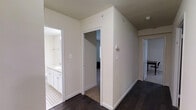 This single-floor residence offers a clean, contemporary aesthetic defined by neutral tones and quality finishes throughout. The interior is anchored by rich, dark hardwood flooring that runs through the main hallway and living areas, providing a striking contrast against the crisp white walls and trim. The hallway serves as the central artery of the home, connecting multiple rooms with an open and airy flow. A well-designed bathroom is accessible directly from the corridor, featuring white cabinetry with ample storage, a vanity with countertop space, and classic tile flooring with a decorative pattern that adds a subtle design detail. The bedroom, visible through an adjoining doorway, continues the dark hardwood flooring and benefits from the home's overall sense of calm and light. White door frames and baseboards provide clean architectural definition throughout, while the neutral wall palette keeps the space feeling bright and versatile. The layout is practical and efficient, with each room thoughtfully positioned for ease of access. Overall, this residence presents a polished, move-in-ready environment with consistent design choices that create a cohesive and inviting atmosphere from room to room.