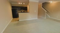 This two-story residence offers a well-designed open-concept layout on the main level, where the kitchen and living area flow seamlessly into one another. The kitchen is equipped with dark espresso cabinetry, black appliances including a refrigerator, microwave, and cooktop, and a dark countertop that contrasts cleanly with the warm beige walls. A peninsula-style breakfast bar separates the kitchen from the living space, providing both a casual dining option and a natural visual boundary between the two areas. The kitchen floor features tile, while the living area transitions to plush wall-to-wall carpeting, adding warmth underfoot. Crown molding lines the upper walls, lending a polished architectural detail throughout the main level. Recessed track lighting in the kitchen ensures the cooking area is well-illuminated. A staircase with white balusters and a wood handrail is visible at the rear of the living space, leading to the second floor. The overall color palette of warm beige tones and dark fixture accents creates a cohesive, neutral aesthetic throughout the main living areas.