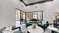 This thoughtfully designed single-floor community space combines sophisticated aesthetics with functional versatility across three interconnected areas. Soaring ceilings with bold dark crown molding create a dramatic sense of vertical openness, while floor-to-ceiling black-framed glass partitions divide the space with architectural elegance, maintaining visual connectivity throughout.

The main lounge area anchors the space with plush emerald green armchairs arranged around a round wood coffee table atop a textured area rug, offering a welcoming and comfortable gathering environment. Large windows along multiple walls flood the interior with abundant natural light and provide views of the surrounding greenery outdoors.

Two dedicated meeting or dining areas branch off from the central lounge, each featuring its own distinct character. One showcases a rustic rectangular wood table surrounded by charcoal upholstered chairs beneath a striking abstract canvas painting. The other offers a round bistro-style table with sleek modern chairs, bathed in natural light from oversized windows. Contemporary abstract artwork adorns the walls throughout, adding curated visual interest.

The entrance corridor features hardwood-style flooring, black-framed glass entry doors with stone exterior accents, and a welcoming accent chair, setting an impressive first impression. The overall ambiance is modern, refined, and inviting — ideal for social gatherings, meetings, or quiet relaxation.