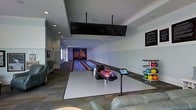 This single-story recreational space is centered around a striking two-lane bowling alley, offering an impressive entertainment venue designed for leisure and social gatherings. The lanes extend toward a vibrant, eye-catching mural at the far end, adding a lively visual focal point to the space. Recessed lighting illuminates the lanes evenly, while a large overhead scoring display is suspended from the ceiling, ensuring visibility for all players.

At the approach area, a ball return system is stocked with a variety of multicolored bowling balls, complemented by a digital scoring console for easy game management. A dedicated ball rack on the side holds additional balls, catering to multiple players simultaneously. The flooring transitions from the polished hardwood lanes to tiled areas, defining the functional zones within the room.

Comfortable seating, including leather sofas, is arranged near the lanes to accommodate spectators and waiting players. The light-toned walls and recessed ceiling details lend the space a clean, modern aesthetic. A window along one wall introduces natural light, balancing the ambient artificial lighting and contributing to an inviting atmosphere throughout this well-appointed recreational area.