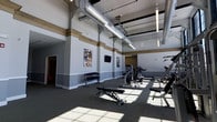 This fitness center is designed to support a comprehensive workout experience within a single, open-plan floor. The space immediately impresses with its soaring ceilings, which are accented by exposed silver ductwork and industrial-style suspended strip lighting — a design choice that lends the room a modern, functional aesthetic. The walls are finished in a two-tone gray palette with warm tan crown molding details, adding a polished character to the otherwise utilitarian setting. Large windows allow natural light to flood across the carpeted floor, creating bright, inviting workout zones throughout the day. The equipment layout is thoughtfully arranged, featuring a dedicated free weights area with a dumbbell rack and adjustable bench, as well as multiple strength training cable and resistance machines positioned to maximize usable floor space. A mounted flat-screen television on the wall provides entertainment for users during their sessions. The open floor plan allows for generous circulation between stations, making the space accessible and easy to navigate. A doorway visible at one end suggests access to additional adjoining areas. Overall, the fitness center strikes a balance between industrial design elements and a clean, contemporary finish, creating a motivating and well-organized environment suited to a wide range of fitness activities.