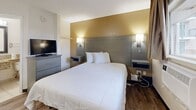 Located in the Siegel community of Bartlett, Tennessee, this single-story unit offers a straightforward, functional layout designed for comfortable everyday living. The bedroom features wood-style flooring, a king-sized bed with a upholstered headboard panel set against a warm gold accent wall, flanked by built-in wall-mounted nightstands and sconce lighting. A wall-mounted flat-screen television and a multi-drawer dresser complete the sleeping area. A window unit provides individual climate control, and a window offers views of the surrounding outdoor greenery.

The kitchen is equipped with white cabinetry, granite-style countertops, a full-size refrigerator, microwave, and a sink, providing essential cooking and storage functionality. Adjacent to the kitchen, a compact dining area accommodates a two-person table with chairs, accented by framed botanical artwork on the wall. The flooring transitions seamlessly between wood-style planks and tile throughout the unit.

The bathroom includes a walk-in shower stall with a curtain, a toilet, a granite-topped vanity, and a towel bar, all finished with neutral tile flooring. The overall aesthetic is clean and practical, reflecting a no-frills, utilitarian design approach.