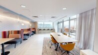 This thoughtfully designed collaborative space offers a bright, contemporary environment suited for work, meetings, and social gatherings. The open-plan layout is organized into distinct functional zones, each contributing to a versatile and welcoming atmosphere. Light wood-look flooring runs throughout, complemented by a neutral color palette that keeps the space feeling airy and open.

At the heart of the main area sits a long communal table surrounded by mustard-yellow upholstered chairs, anchored by a woven area rug that adds warmth and texture. Flanking one wall is a row of intimate booth-style seating — colorful striped high-back banquettes paired with small café tables — ideal for casual conversations or focused work. Above, accent lighting and a geometric-patterned wall treatment add visual interest and depth.

A built-in bar-height counter along the windowed wall provides additional seating with bar stools, offering an elevated workspace bathed in natural light. Full-height glazed doors open directly onto a furnished outdoor terrace adorned with murals, extending the usable area into the open air with city views beyond.

A private room off the main space features a work table, built-in desk, and sheer drapery, functioning as a quieter breakout area. A drop ceiling with recessed lighting ensures consistent, well-distributed illumination throughout.