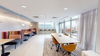 This thoughtfully designed collaborative space offers a bright, contemporary environment suited for work, meetings, and social gatherings. The open-plan layout is organized into distinct functional zones, each contributing to a versatile and welcoming atmosphere. Light wood-look flooring runs throughout, complemented by a neutral color palette that keeps the space feeling airy and open.

At the heart of the main area sits a long communal table surrounded by mustard-yellow upholstered chairs, anchored by a woven area rug that adds warmth and texture. Flanking one wall is a row of intimate booth-style seating — colorful striped high-back banquettes paired with small café tables — ideal for casual conversations or focused work. Above, accent lighting and a geometric-patterned wall treatment add visual interest and depth.

A built-in bar-height counter along the windowed wall provides additional seating with bar stools, offering an elevated workspace bathed in natural light. Full-height glazed doors open directly onto a furnished outdoor terrace adorned with murals, extending the usable area into the open air with city views beyond.

A private room off the main space features a work table, built-in desk, and sheer drapery, functioning as a quieter breakout area. A drop ceiling with recessed lighting ensures consistent, well-distributed illumination throughout.