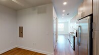 This single-floor unit offers a thoughtfully designed layout with modern finishes throughout. The kitchen is a standout feature, equipped with warm wood-toned cabinetry extending to the ceiling, light quartz countertops, and a full suite of stainless steel appliances including a refrigerator, range, microwave, and dishwasher. Recessed lighting illuminates the space, which connects seamlessly to the main living area via an open corridor. The living and utility areas feature warm hardwood-style flooring that runs continuously throughout the unit, contributing to a cohesive aesthetic. A dedicated utility space includes an electrical panel and HVAC venting, providing practical infrastructure. The bedroom is bright and airy, benefiting from a wide, multi-pane window that frames outdoor greenery and allows generous natural light. A ceiling fan with integrated lighting adds comfort and functionality. The bathroom is well-appointed with a wood-grain floating vanity, white quartz countertop, an undermount sink, and a backlit mirror that provides even, flattering illumination. A full soaking tub with a tiled surround completes the bathroom. Clean white walls and recessed lighting throughout reinforce the unit's modern, minimal character.