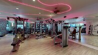 This single-floor fitness facility is designed to accommodate a comprehensive range of workout needs within a thoughtfully organized layout. The space is immediately distinguished by its signature pink neon lighting, which traces fluid, wave-like patterns across the white ceiling, creating an energetic yet stylish ambiance throughout. Ceiling fans are positioned at intervals to maintain airflow across the workout floor.

The main strength training area is generously proportioned, featuring an extensive array of Life Fitness cable and weight machines arranged in rows, allowing for efficient use of space and easy navigation between equipment. Full-length mirrors line multiple walls, visually expanding the room while providing form-checking functionality. A dedicated free weights zone includes a well-stocked dumbbell rack alongside adjustable benches and kettle bells, catering to both beginner and advanced users.

A separate cardio section houses treadmills and elliptical machines, positioned near windows that allow natural light to enter and offer exterior views. A wall-mounted flat-screen television is installed in this area for entertainment during workouts. The flooring throughout is a light-toned wood-style surface that is both durable and visually clean. A textured geometric accent wall panel adds a decorative element near the cardio zone. An entry area with glass-panel doors provides access to a reception or office space.