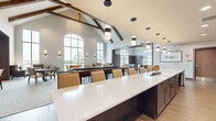 This expansive club room in Menomonee Falls, WI, offers a thoughtfully designed, single-floor community gathering space that blends upscale amenities with a warm, inviting atmosphere. The architectural centerpiece is the dramatic vaulted ceiling adorned with exposed wood beams, lending a refined yet rustic character to the space. Large arched and floor-to-ceiling windows flood the interior with abundant natural light while offering views of the surrounding neighborhood.

The club room is organized into several distinct functional areas. A full-length bar with built-in shelving, pendant lighting, and bar-height seating serves as a social hub. Adjacent to the bar, a regulation shuffleboard table anchors the game area, complemented by multiple wall-mounted televisions. A dedicated billiards room features rich wood panel wainscoting, coffered ceilings with exposed beam accents, and ample natural light from arched windows.

The dining and lounge zones are furnished with a mix of upholstered chairs, armchairs, and dining tables, creating flexible seating arrangements for both casual gatherings and structured dining. Hardwood and carpet flooring define different zones throughout, while warm, layered lighting — including recessed fixtures, pendant lights, and wall sconces — contributes to a comfortable and sophisticated ambiance throughout this generously sized community space.