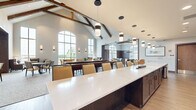 This expansive club room in Menomonee Falls, WI, offers a thoughtfully designed, single-floor community gathering space that blends upscale amenities with a warm, inviting atmosphere. The architectural centerpiece is the dramatic vaulted ceiling adorned with exposed wood beams, lending a refined yet rustic character to the space. Large arched and floor-to-ceiling windows flood the interior with abundant natural light while offering views of the surrounding neighborhood.

The club room is organized into several distinct functional areas. A full-length bar with built-in shelving, pendant lighting, and bar-height seating serves as a social hub. Adjacent to the bar, a regulation shuffleboard table anchors the game area, complemented by multiple wall-mounted televisions. A dedicated billiards room features rich wood panel wainscoting, coffered ceilings with exposed beam accents, and ample natural light from arched windows.

The dining and lounge zones are furnished with a mix of upholstered chairs, armchairs, and dining tables, creating flexible seating arrangements for both casual gatherings and structured dining. Hardwood and carpet flooring define different zones throughout, while warm, layered lighting — including recessed fixtures, pendant lights, and wall sconces — contributes to a comfortable and sophisticated ambiance throughout this generously sized community space.