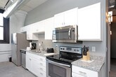 This single-floor loft-style space masterfully combines industrial heritage with modern comfort. The open layout is anchored by striking exposed white-painted brick walls and soaring ceilings with visible ductwork and track lighting, reflecting the building's historic character throughout.

The kitchen is well-appointed with white shaker cabinetry, granite countertops, and a full suite of stainless steel appliances including a range, dishwasher, microwave, and refrigerator, all set against polished concrete flooring. Black-framed windows throughout the space flood each room with natural light while offering urban views.

The living area features the same exposed brick backdrop, creating a warm, loft-like ambiance ideal for relaxing or entertaining. Two bedrooms are present: one showcases the original brick walls and benefits from natural light through a black-trimmed window, while the second features smooth painted walls, wall sconce lighting, and a double-door closet. Both rooms are comfortably appointed.

A full bathroom with tile flooring, a single-sink vanity, and a shower-tub combination completes the layout. The consistent industrial aesthetic, marked by concrete floors, exposed infrastructure, and historic masonry, gives this space a distinctive and cohesive character.