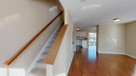 This three-floor property in Hamburg offers a practical and well-structured layout with consistent warm beige tones throughout. The main living area features rich hardwood flooring that extends toward an open-plan kitchen, which is connected to the living space via a half-wall breakfast bar with a wooden ledge. The kitchen is equipped with white cabinetry, light countertops, stainless steel appliances including a range, built-in microwave, and a double stainless steel sink, providing ample storage and functionality. A carpeted staircase with a wooden handrail connects the floors, contributing to the home's cohesive aesthetic. The upper floors host multiple generously sized bedrooms, each finished with plush carpeting and fitted with multiple windows that allow abundant natural light to fill the rooms. The bathrooms include one with a full soaking tub and shower combination, and another featuring a standalone walk-in shower enclosed with frosted glass sliding doors, both finished with tile flooring. A dedicated laundry room with side-by-side washer and dryer units, wire shelving, and a window rounds out the home's practical amenities.