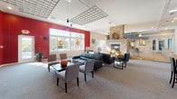 Located at 1 Apple Rd in Beverly, MA, this single-floor commercial space offers a welcoming and well-appointed interior designed for professional use. The centerpiece of the main area is a striking floor-to-ceiling stacked stone fireplace that anchors the open layout and adds warmth and character to the environment. Bold red accent walls contrast with crisp white ceilings and neutral carpeting, creating a dynamic yet professional atmosphere throughout.

The main lounge area is generously proportioned, featuring multiple seating arrangements including leather sofas, armchairs, and dining-height tables with chairs—accommodating both casual meetings and more structured gatherings. A wall-mounted flat-screen TV and built-in speakers enhance the functionality of the space. Pendant lighting with deep blue glass shades hangs from a decorative lattice ceiling element, adding a refined architectural detail overhead.

A tiled entryway with a reception counter provides a clean, professional welcome point upon arrival. The kitchenette is equipped with wood cabinetry, dark granite countertops, and modern appliances, with a window offering views of an outdoor patio area. Large windows along the perimeter flood the space with natural light and provide pleasant outdoor views, contributing to an open and inviting overall ambiance.
