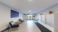 The Blueprint Residences presents a thoughtfully designed common area layout across a single floor, encompassing a generously sized lobby, connecting hallways, and a resident lounge area. The main lobby serves as the central welcoming space, featuring light grey tile flooring, clean white walls with soft blue accents, and recessed lighting that creates a bright, professional atmosphere. A bold branded sign prominently displays "The Blueprint Residences," anchoring the space with a strong identity. The lobby is furnished with contemporary black leather seating and a round wooden side table, offering a comfortable waiting area for residents and guests. Natural light streams in through the glass entry doors, providing views to the exterior. A comprehensive bank of individual resident mailboxes lines one wall, accompanied by recycling stations and clearly posted fire safety information, ensuring practical functionality alongside the modern aesthetic. The hallway corridors extend from the lobby, finished with patterned commercial-grade carpeting, recessed ceiling lighting, and crisp white walls that lead residents efficiently to their units. The overall ambiance throughout is clean, contemporary, and functional, reflecting a well-maintained residential building designed to balance practicality with modern design sensibilities.