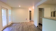 This single-floor space offers a bright and functional layout designed with modern, neutral finishes throughout. The main living area features warm, wood-style laminate flooring that extends through the space, complemented by smooth white walls and recessed ceiling lighting that create a clean, well-lit ambiance. A dedicated hallway connects the living area to the additional rooms beyond, providing a clear and practical flow through the unit. A built-in closet with shelving is visible off the main living area, offering convenient and organized storage. The kitchen area is partially visible with light-toned cabinetry and what appears to be a granite or stone-style countertop, suggesting thoughtful material choices throughout. The overall design is minimal and move-in ready, with neutral tones that lend themselves well to a variety of interior styles. Recessed and flush-mount ceiling fixtures provide consistent illumination throughout. The space is characterized by its simplicity, practicality, and cohesive aesthetic, making it a versatile and comfortable single-level living environment.