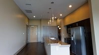 This contemporary single-floor residence showcases a thoughtfully designed open-concept layout that seamlessly blends functionality with modern aesthetics. The heart of the space is the expansive kitchen, anchored by a striking center island finished in crisp white cabinetry and topped with polished granite countertops that catch the light beautifully. A sleek, modern faucet and integrated sink add to the island's practicality.

Overhead, a cluster of elegant glass pendant lights hangs at varying lengths, serving as a focal point and providing warm, ambient illumination. The kitchen is further equipped with a full suite of stainless steel appliances, including a refrigerator, microwave, and range, all complemented by rich, dark upper cabinetry that adds depth and contrast to the space.

The flooring throughout is rich dark hardwood, lending warmth and sophistication to the open floor plan. Neutral gray walls provide a versatile backdrop, while recessed lighting supplements the pendant fixtures to ensure the space is well-lit throughout. A natural wood-toned entry door adds an organic touch, offering a welcoming contrast to the sleek, contemporary finishes found throughout the rest of the space.