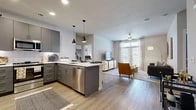 This modern single-floor residence offers a well-designed layout featuring one bedroom, two full bathrooms, a dedicated office, and two closets, making efficient use of its generous floor area. The open-concept kitchen and living area form the heart of the home, anchored by wide-plank hardwood-style flooring that flows seamlessly throughout. The kitchen is outfitted with slate-gray cabinetry, stainless steel appliances including a French door refrigerator and dishwasher, a bright white quartz peninsula with bar seating for three, and stylish pendant lighting overhead. The adjoining living room benefits from large windows dressed with sheer curtains, filling the space with abundant natural light. The bedroom features plush carpet, generous natural light from large windows, and access to a walk-in closet with built-in shelving and hanging rods. The bathrooms echo the kitchen's modern aesthetic with matching gray cabinetry, white countertops, and tile-surround tub-shower combinations. The dedicated office is carpeted and bright, with a large window and ample space for a full desk setup. Throughout the residence, a neutral color palette and consistent modern finishes create a cohesive, contemporary ambiance.