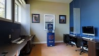 This Auburn University business center is a compact, single-floor facility designed to serve students and visitors with essential computing and printing resources. The space is immediately recognizable by its Auburn University branding, most notably a prominent blue self-service "Fan Connection" printing kiosk displayed at the center of the room, featuring the iconic Auburn "AU" logo. The walls are painted in a warm beige tone, accented by deep navy blue partitions and trim that reinforce the school's signature colors. Framed photographs of Auburn University campus landmarks adorn the walls, adding a sense of institutional pride and character to the environment. The flooring consists of warm terracotta-toned tile, lending the space a grounded, practical aesthetic. A computer workstation with a monitor and ergonomic chair is positioned along the right side of the room, while additional desk space along the left wall accommodates further work needs. Natural light filters in through a centrally placed window, brightening the interior and creating a welcoming atmosphere. A pass-through window suggests a connection to an adjacent service or reception area. Overall, the space is efficiently laid out, clean, and purpose-driven — providing a convenient, on-brand resource center for the Auburn University community.