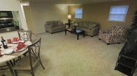 This single-floor space offers a comfortable and functional layout designed for everyday living. The living area serves as the centerpiece of the home, showcasing a generous, open floor plan that provides ample room for relaxation and entertaining. Soft, neutral-toned carpeting extends throughout the space, contributing to a warm and inviting atmosphere. The room is furnished with a coordinated seating arrangement, including two sofas and an accent chair, all in earthy tones and complemented by decorative throw pillows that add subtle visual interest. A dark wood coffee table and matching side table anchor the seating area, while a table lamp provides warm ambient lighting. The walls are finished in a muted olive-green hue, lending a cozy, understated character to the space. A window fitted with blinds allows natural light to filter into the room, brightening the interior. A decorative chair visible at the edge of the space hints at an adjacent dining or multi-use area, suggesting a versatile and well-considered floor plan. Overall, the space conveys a sense of ease and practicality, ideal for comfortable, day-to-day living.