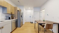 Located at 399 Crouse Ave in Syracuse, NY, this thoughtfully designed studio offers a bright, open-concept layout that efficiently combines living and sleeping areas on a single floor. Light-toned hardwood flooring flows throughout the entire space, lending a clean, cohesive aesthetic.

The kitchen is well-appointed with stainless steel appliances — including a French door refrigerator with ice maker, electric range, over-the-range microwave, and dishwasher. Two-tone cabinetry pairs natural wood upper cabinets with sleek grey lower units, complemented by light countertops and a single-basin sink. A large window floods the kitchen with natural light.

The open living area provides comfortable space for both lounging and sleeping, with a sliding barn-style door adding a modern architectural touch. Recessed lighting and large windows maintain a bright, airy ambiance throughout.

The dedicated walk-in closet offers generous storage with wire shelving systems and multiple hanging rods along all walls. The bathroom features a grey vanity with a white countertop, matching the overall modern palette, and continues the light hardwood flooring. A keypad-secured entry door adds a contemporary convenience to this well-designed urban residence.