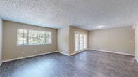This single-floor residence offers a clean, functional layout designed for comfortable everyday living. The main living area is a generously sized open space finished with durable gray wood-look flooring and neutral warm-beige walls, complemented by a textured popcorn ceiling. A large picture window floods the room with natural light and frames views of lush greenery outside, while a set of white-framed French doors provides an additional connection to the outdoors, enhancing the bright and airy feel of the space.

The bedrooms are carpeted in soft gray, offering a cozy and quiet retreat. Each room features neutral walls, white panel closet doors, and windows fitted with blinds that allow for natural light while maintaining privacy. The rooms are well-proportioned, providing ample space for furnishings.

The bathroom is finished in a crisp, all-white palette, featuring a full soaking tub with subway tile surround, a white vanity with a light stone countertop, and a mirrored medicine cabinet above. The flooring transitions to wood-look tile, maintaining a cohesive aesthetic throughout.

A dedicated in-unit laundry room houses a full-size top-loading washer and dryer, adding a layer of everyday convenience. Throughout the home, clean white baseboards, consistent neutral tones, and well-maintained surfaces create a move-in-ready atmosphere.