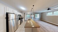 This single-floor residence offers a thoughtfully designed open-concept layout that seamlessly connects the kitchen, dining, and living areas. The kitchen is well-equipped with stainless steel appliances, including a refrigerator, electric range, and microwave, all complemented by crisp white cabinetry, dark granite countertops, and light ceramic tile flooring. A granite-topped island with a built-in sink and pendant lighting serves as the centerpiece of the space, providing both functionality and style. The adjoining living area features plush wall-to-wall carpeting, neutral gray walls, and multiple windows that invite an abundance of natural light and offer views of surrounding greenery. A ceiling fan adds comfort to the living space, which also provides access to an outdoor area. The bedrooms are generously sized, each finished with soft carpeting, ceiling fans, and windows framing lush outdoor views, creating a calm and comfortable retreat. Both bathrooms are finished with granite-topped vanities, tile flooring, and oval mirrors with accent lighting. The primary bathroom includes a walk-in closet area and a tub-shower combination, while the secondary bathroom mirrors a similar clean, functional design. Overall, the space presents a cohesive, neutral palette throughout.