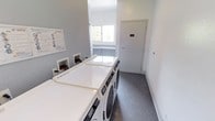 This shared laundry facility is designed with practicality and efficiency in mind. The room is outfitted with a row of commercial-grade front-loading washing machines and dryers positioned along one wall, providing ample capacity for resident use. A built-in folding surface sits atop the machines, offering a convenient space for sorting and folding laundry. The walls are finished in a cool grey tone, contributing to the room's clean and modern aesthetic, while dark-toned flooring adds a grounded contrast. A window at the far end of the room allows natural light to fill the space, creating a bright and welcoming environment. Posted instructions and informational signage are mounted on the wall near the machines, ensuring ease of use for all occupants. A clearly labeled door provides access to an adjacent utility or storage area. The overall layout is thoughtfully organized to maximize usability, with open floor space allowing for comfortable movement throughout the room. This well-maintained laundry room reflects a commitment to providing a clean, functional, and accessible shared amenity.