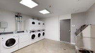 This single-floor shared laundry facility is designed with functionality and cleanliness in mind. The space is finished with smooth concrete flooring that is both durable and easy to maintain, providing a practical foundation for a high-use utility area. Bright flush-mounted panel lighting is installed across the ceiling, ensuring the room is well-illuminated throughout.

The laundry room is equipped with multiple commercial-grade front-loading washing machines and stacked dryer units, all finished in white and arranged neatly along the wall. The machines offer users a full suite of washing and drying options. The walls are painted in a crisp, neutral white, contributing to the overall clean and modern aesthetic of the space.

A solid gray access door is situated along one wall, providing a clear entry and exit point. The room maintains a spacious, open feel, allowing users to move comfortably between machines. The minimalist design keeps the focus on utility, with no unnecessary fixtures or furnishings cluttering the area. Overall, this laundry facility presents a well-organized, hygienic, and efficiently designed space suited for shared or communal use.
