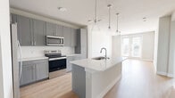 This well-designed 1-bedroom, 1-bathroom residence offers a thoughtful, single-level layout that maximizes functionality and modern style. The open-concept kitchen and living area serves as the heart of the home, featuring light hardwood flooring, gray shaker-style cabinetry, white quartz countertops, and a full suite of stainless steel appliances including a refrigerator with ice maker, electric range, and over-the-range microwave. A central island with an undermount sink and pendant lighting above provides both additional workspace and a natural gathering point. French doors in the living area invite natural light and offer outdoor access, complemented by a ceiling fan overhead. The bedroom is a comfortable, carpeted retreat bathed in natural light from dual windows with views of the surrounding cityscape, and is equipped with its own ceiling fan. The full bathroom is appointed with gray vanity cabinetry, white countertops, a large framed mirror, and a soaking tub with tiled surround, with direct access to a dedicated closet featuring built-in shelving. A hallway connects the spaces cohesively, and the overall neutral color palette of whites and warm grays creates a clean, contemporary ambiance throughout.