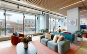 This contemporary coworking space is defined by its striking architectural details and thoughtfully curated interiors. A signature slatted wood ceiling runs throughout much of the floor, lending warmth and visual depth while integrated linear lighting ensures a well-lit environment. Light hardwood flooring unifies the space and complements the vibrant color palette found in every zone.

The expansive lounge area is anchored by floor-to-ceiling windows that flood the space with natural light and offer sweeping views of the surrounding urban streetscape. Plush seating arrangements — including teal sofas, rust-toned chairs, and a sculptural coffee table atop a terracotta rug — create an inviting, residential-inspired atmosphere. Built-in shelving units curated with books, plants, and art add a refined, personalized touch along corridor walls.

A generously sized café-style dining area provides multiple seating configurations, from marble-topped counter seating with arc pendant lighting to communal wooden tables, all bathed in natural light from large windows and glass doors.

Glass-partitioned meeting rooms lined along a corridor offer transparency and privacy in equal measure, with bold green and navy accent walls adding character. A fully equipped kitchen — outfitted with bold red cabinetry, a built-in oven, induction cooktop, and tiled backsplash — serves the space with both style and function. Throughout, curated artwork and lush greenery reinforce a creative, energetic ambiance.