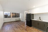 Located at 1060 Bishop St in the heart of Honolulu, HI, this 2-bedroom, 1-bathroom unit offers a practical and modern layout across a single floor. The kitchen is outfitted with contemporary gray cabinetry, white countertops, a black refrigerator, electric range, and recessed lighting — all set over warm wood-look vinyl flooring that extends into the living area. Large windows in the living space frame views of downtown Honolulu's urban skyline, allowing abundant natural light to fill the room. Each bedroom is carpeted for comfort and features its own closet with sliding doors. The bedrooms are equipped with wall-mounted PTAC air conditioning units and windows that bring in additional natural light and city views. The bathroom presents a clean, modern aesthetic with a floating vanity finished in a warm taupe tone, white quartz countertop, undermount sink, and matte black fixtures, complemented by a large frameless mirror. Tile flooring in the bathroom contrasts neatly with the wood-look flooring found throughout the rest of the unit. The overall design is clean and contemporary, with a neutral palette of whites and grays creating a bright, cohesive atmosphere throughout.