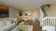 This two-story residence offers a thoughtfully designed layout that maximizes both comfort and functionality. The main level features an open-concept floor plan where the living area, dining space, and kitchen flow seamlessly into one another. Rich, dark hardwood flooring runs throughout, providing a warm and cohesive foundation for the entire level.

The living area is bright and inviting, with neutral taupe-toned walls that create a versatile backdrop for a range of furnishing styles. A glass-topped coffee table and a plush sofa anchor the space, while decorative accents add character and warmth. Adjacent to the living area, the dining space accommodates a full dining set with white lattice-back chairs, positioned beneath modern track lighting that provides both ambient and task illumination.

The kitchen is accessible directly from the dining area and features a pass-through window that enhances the open feel of the main level. A hallway leads toward additional rooms and a closeted entry, while a white-railed staircase with carpeted treads provides access to the upper floor. The overall ambiance of the home is clean, airy, and move-in ready, with a neutral color palette and classic architectural details that lend timeless appeal throughout.