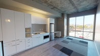 This contemporary single-floor studio showcases an industrial-modern aesthetic defined by its signature exposed concrete ceiling, which adds raw texture and visual character throughout the open-plan layout. The living and kitchen areas flow seamlessly together, creating a versatile and efficient use of space.

The kitchen is thoughtfully designed with crisp white flat-front cabinetry extending from floor to ceiling, providing generous storage. Stainless steel appliances — including a full-size refrigerator and built-in microwave — complement the clean, minimalist aesthetic. A single-basin sink and integrated cooktop complete the functional kitchenette setup. A built-in desk or counter extension runs adjacent to the kitchen, offering a dedicated workspace or dining area.

The main living space features a combination of light hardwood-style flooring and modular carpet tile inlays, adding warmth and visual interest underfoot. A bold red entry door creates a striking focal point at the entrance, contrasting with the otherwise neutral palette of soft grays and whites. Wall sconces near the entry provide warm accent lighting.

Large sliding glass doors along one wall flood the interior with natural light and open to an outdoor view of the surrounding cityscape, enhancing the sense of openness and connection to the urban environment. Recessed linear lighting supplements the natural light for a well-lit, inviting atmosphere throughout.