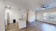 This single-floor residence offers a clean, modern aesthetic defined by an open-concept design and consistent hardwood-style flooring throughout. The living area is bright and spacious, anchored by a ceiling fan with light fixture and a large window that invites ample natural light and outdoor views. It flows seamlessly into the kitchen, which features a central island with granite countertops and a matte black pull-down faucet, white cabinetry, and stainless steel appliances including a refrigerator, range, and over-the-range microwave. A built-in desk nook with matching granite surface provides a convenient workspace within the main living area. Track lighting illuminates key zones, adding to the modern, well-lit ambiance. The bedroom is generously proportioned with light-toned hardwood-style flooring, crisp white walls, and a window offering natural light. The bathroom is finished with white square tile surrounding a full tub-shower combination, complemented by oil-rubbed bronze fixtures and hardware. Throughout the space, white walls and clean lines create a cohesive, contemporary environment that is both functional and inviting.
