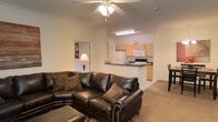 This single-floor residence showcases an open-concept layout that seamlessly connects the living area to the kitchen. The living room is anchored by a substantial dark leather sectional sofa accented with decorative pillows, complemented by a matching ottoman. Crown molding runs along the ceiling perimeter, adding a refined architectural detail, while a ceiling fan with integrated lighting provides both comfort and illumination. Neutral-toned walls and light carpeting create a warm, cohesive ambiance throughout the space. The kitchen is positioned adjacent to the living area and is separated by a peninsula breakfast bar featuring a dark countertop that provides additional seating and prep space. Light wood cabinetry lines the kitchen walls, offering ample storage, while white appliances — including a refrigerator, microwave, and range — maintain a clean, consistent aesthetic. Recessed and overhead fluorescent lighting keeps the kitchen well-lit. A doorway visible from the living area suggests additional rooms beyond, contributing to the overall functional flow of the residence.