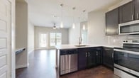 This single-floor residence presents a thoughtfully designed open-concept layout that seamlessly connects the kitchen and living areas. The kitchen serves as a central focal point, featuring dark espresso cabinetry with sleek hardware, crisp white quartz countertops, and a peninsula with an undermount sink. A full suite of stainless steel appliances — including a refrigerator, range, microwave, and dishwasher — is complemented by a classic subway tile backsplash. Three pendant lights hang above the peninsula, providing focused task lighting, while a ceiling fan overhead serves the adjacent living area. French doors in the living space allow natural light to fill the room and provide exterior access, with soft carpet underfoot adding warmth and comfort. Rich, dark hardwood-style flooring flows throughout the kitchen and main areas, creating a striking contrast with the neutral wall tones. Additional rooms feature hardwood-style or carpeted floors and are well-lit with natural light from windows with blinds. The bathroom is finished with a frameless glass-enclosed shower, dark vanity cabinetry, and coordinating hardwood-style flooring, maintaining a consistent modern aesthetic throughout the unit.