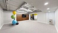 Located at 5764 Stevens Forest Rd in Columbia, Maryland, this fitness studio offers a thoughtfully designed workout space that balances functionality with modern style. The space is organized into two distinct zones: an open, carpeted exercise floor ideal for stretching, yoga, and functional training, and an adjacent equipment area finished with durable rubber flooring that houses strength machines, a cable system, and cardio equipment including treadmills and a stationary bike.

The open exercise area serves as the heart of the studio, featuring a striking slatted wood accent wall with a wall-mounted flat-screen TV — perfect for guided workouts or entertainment during training sessions. A well-stocked equipment rack lines one wall, holding an assortment of stability balls, medicine balls, resistance bands, and foam rollers to support a wide variety of fitness routines.

The equipment zone, distinguished by its bold yellow-painted ceiling structure, adds an energetic visual contrast to the otherwise neutral palette of white walls and gray flooring. Overhead, an exposed ceiling with industrial-style fluorescent linear lighting and visible ductwork gives the space a raw, utilitarian character while ensuring bright, even illumination throughout. The layout flows openly between zones, making the studio versatile and easy to navigate for a full-body workout experience.