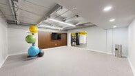 Located at 5764 Stevens Forest Rd in Columbia, Maryland, this fitness studio offers a thoughtfully designed workout space that balances functionality with modern style. The space is organized into two distinct zones: an open, carpeted exercise floor ideal for stretching, yoga, and functional training, and an adjacent equipment area finished with durable rubber flooring that houses strength machines, a cable system, and cardio equipment including treadmills and a stationary bike.

The open exercise area serves as the heart of the studio, featuring a striking slatted wood accent wall with a wall-mounted flat-screen TV — perfect for guided workouts or entertainment during training sessions. A well-stocked equipment rack lines one wall, holding an assortment of stability balls, medicine balls, resistance bands, and foam rollers to support a wide variety of fitness routines.

The equipment zone, distinguished by its bold yellow-painted ceiling structure, adds an energetic visual contrast to the otherwise neutral palette of white walls and gray flooring. Overhead, an exposed ceiling with industrial-style fluorescent linear lighting and visible ductwork gives the space a raw, utilitarian character while ensuring bright, even illumination throughout. The layout flows openly between zones, making the studio versatile and easy to navigate for a full-body workout experience.