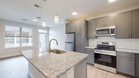 This thoughtfully designed one-bedroom, one-bathroom residence in Rochester, MN offers a modern and functional layout across a single floor. The kitchen serves as the centerpiece of the space, showcasing granite countertops, a large island with an undermount sink and pendant lighting, gray shaker-style cabinetry, a white subway tile backsplash, and a full suite of stainless steel appliances including a refrigerator, electric range, and over-the-range microwave. Light wood-look flooring runs throughout the main living areas, enhancing the bright, clean aesthetic. A hallway connects the living area to the private bedroom, which features plush wall-to-wall carpet and neutral tones that create a calm, restful atmosphere. The dedicated closet provides practical storage solutions. The bathroom is finished with a clean, all-white tub and shower combination featuring brushed nickel fixtures, complemented by light wood-look flooring that ties the space together. Recessed lighting and large windows allow natural light to flow throughout, contributing to an open and airy feel. The overall design balances contemporary style with everyday practicality, making this a comfortable and attractive residence.