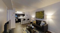 This one-bedroom, one-bathroom apartment is thoughtfully designed to maximize every square foot of its compact footprint. The open-concept layout connects the living area and kitchen, creating a cohesive and functional living space. The living room features wood-style flooring, neutral walls, and a textured ceiling, providing a warm and inviting atmosphere. A comfortable seating area anchors the room, complemented by accent artwork and stylish décor that add a pop of color and personality. The kitchen is equipped with dark cabinetry, black appliances, and a light-toned countertop that contrasts boldly with the rich cabinet finish. A breakfast bar with seating for two serves as both a dining and casual gathering spot, further enhancing the open feel of the space. The flooring flows seamlessly from the living area into the kitchen, unifying the two zones visually. The layout is efficient and well-organized, making the most of the available space without sacrificing comfort or style. Overall, this apartment offers a modern, practical interior with a cohesive design aesthetic suited for individuals or couples seeking a well-appointed living space.