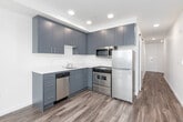 This single-floor unit showcases a thoughtfully designed open layout with a consistent palette of crisp white walls and warm wood-tone flooring throughout. The kitchen is a functional highlight, featuring two-tone cabinetry with charcoal upper cabinets and muted blue-grey lower cabinets, white quartz countertops, a tile backsplash, and a full suite of stainless steel appliances including a range, over-range microwave, dishwasher, and refrigerator. The living and sleeping areas benefit from generous natural light delivered through large floor-to-ceiling sliding glass doors and additional operable windows, which frame elevated views of the surrounding cityscape and a glimpse of the waterfront beyond. A sliding barn door adds a contemporary architectural touch to the hallway, which also houses a dedicated laundry closet fitted with a stacked washer and dryer unit. The bathroom is finished with clean, minimalist details, including a white vanity countertop with an undermount sink, a frameless mirror, and a toilet. The overall ambiance of the space is modern and understated, with an emphasis on natural light, urban views, and efficient use of space.