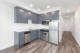 This single-floor unit showcases a thoughtfully designed open layout with a consistent palette of crisp white walls and warm wood-tone flooring throughout. The kitchen is a functional highlight, featuring two-tone cabinetry with charcoal upper cabinets and muted blue-grey lower cabinets, white quartz countertops, a tile backsplash, and a full suite of stainless steel appliances including a range, over-range microwave, dishwasher, and refrigerator. The living and sleeping areas benefit from generous natural light delivered through large floor-to-ceiling sliding glass doors and additional operable windows, which frame elevated views of the surrounding cityscape and a glimpse of the waterfront beyond. A sliding barn door adds a contemporary architectural touch to the hallway, which also houses a dedicated laundry closet fitted with a stacked washer and dryer unit. The bathroom is finished with clean, minimalist details, including a white vanity countertop with an undermount sink, a frameless mirror, and a toilet. The overall ambiance of the space is modern and understated, with an emphasis on natural light, urban views, and efficient use of space.